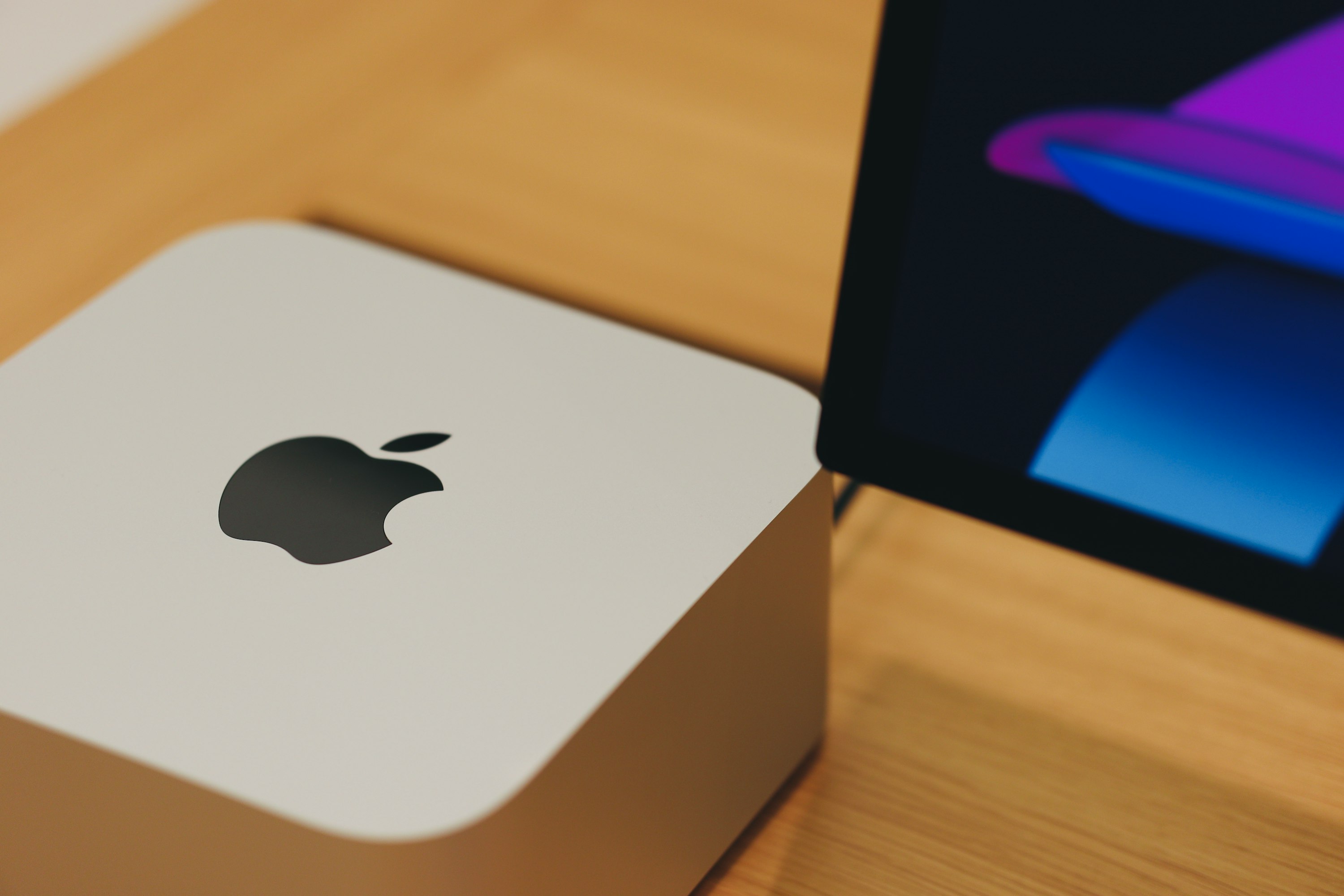 Mac Studio sobre uma superfície de madeira e à esquerda do ecrã de um iMac com apenas o canto inferior esquerdo visível.