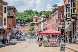 A small-town street lined with historic brick buildings, featuring storefronts with signs and American flags. A red canopy covers outdoor seating, and people are visible walking and interacting. The street is partially closed off with barriers, and cars are parked along the sides. Lush green trees are visible in the background under a partly cloudy sky.