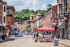 A small-town street lined with historic brick buildings, featuring storefronts with signs and American flags. A red canopy covers outdoor seating, and people are visible walking and interacting. The street is partially closed off with barriers, and cars are parked along the sides. Lush green trees are visible in the background under a partly cloudy sky.