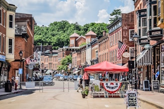 A small-town street lined with historic brick buildings, featuring storefronts with signs and American flags. A red canopy covers outdoor seating, and people are visible walking and interacting. The street is partially closed off with barriers, and cars are parked along the sides. Lush green trees are visible in the background under a partly cloudy sky.