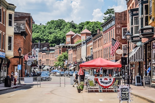 A small-town street lined with historic brick buildings, featuring storefronts with signs and American flags. A red canopy covers outdoor seating, and people are visible walking and interacting. The street is partially closed off with barriers, and cars are parked along the sides. Lush green trees are visible in the background under a partly cloudy sky.