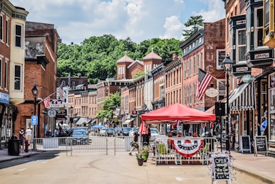 A small-town street lined with historic brick buildings, featuring storefronts with signs and American flags. A red canopy covers outdoor seating, and people are visible walking and interacting. The street is partially closed off with barriers, and cars are parked along the sides. Lush green trees are visible in the background under a partly cloudy sky.