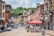 A small-town street lined with historic brick buildings, featuring storefronts with signs and American flags. A red canopy covers outdoor seating, and people are visible walking and interacting. The street is partially closed off with barriers, and cars are parked along the sides. Lush green trees are visible in the background under a partly cloudy sky.