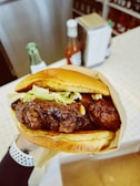 A close-up of a fried chicken sandwich held in a person's hand. The sandwich consists of crispy fried chicken pieces topped with shredded lettuce, nestled in a golden brioche bun. In the background, there is a blurred view of a diner-like counter, with condiments including a ketchup bottle and a napkin holder.
