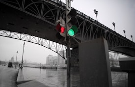 A traffic light displaying green and red signals, positioned beneath a large, intricately designed metal bridge. The background features an urban setting with buildings and a riverbank walkway. The scene is overcast, with wet pavement indicating recent rain.