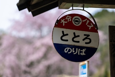 A circular sign with Japanese text is hanging under a roof. The sign is divided into three horizontal sections colored red, white, and blue. In the background, there is a blurred view of trees with pink blossoms, indicating cherry blossoms in a natural setting.