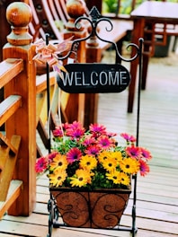 A decorative black wrought iron stand holds a small rectangular sign with the word 'WELCOME' written in white letters. Below the sign, a basket filled with vibrant flowers, primarily in shades of pink and yellow, sits on a wooden porch. The background features parts of a wooden fence and outdoor furniture, contributing to a warm, inviting atmosphere.