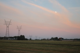 A rural landscape features tall electricity pylons stretching across open fields under a vast sky with soft pastel hues of pink and orange. The moon is visible in the sky, adding a serene touch to the scene. Trees and farmhouses are scattered in the distance, contributing to the peaceful rural setting.