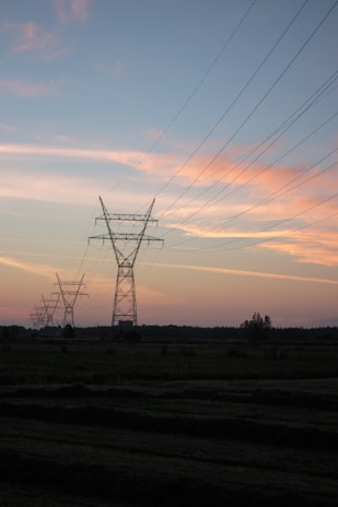 Several transmission towers and power lines stretch across a rural landscape at dusk. The sky displays a gradient of colors from soft pinks and oranges to darker blues, suggesting the setting sun. The land below appears flat with traces of vegetation, silhouetted against the sky.