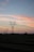 Several transmission towers and power lines stretch across a rural landscape at dusk. The sky displays a gradient of colors from soft pinks and oranges to darker blues, suggesting the setting sun. The land below appears flat with traces of vegetation, silhouetted against the sky.