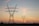 A series of large electricity pylons stretch into the distance against a backdrop of a colorful sunset, with hues of orange, pink, and purple blending into the sky. The landscape is flat, with some greenery visible at the base of the pylons.