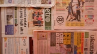 Multiple newspapers are laid out with various headlines and images. The layout includes different sections and pages, with colors ranging from a light tan to vibrant reds and greens. Advertisements and articles are visible with bold headlines and images related to sports and news events. The newspapers are arranged horizontally across a flat surface.