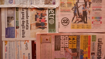 Multiple newspapers are laid out with various headlines and images. The layout includes different sections and pages, with colors ranging from a light tan to vibrant reds and greens. Advertisements and articles are visible with bold headlines and images related to sports and news events. The newspapers are arranged horizontally across a flat surface.