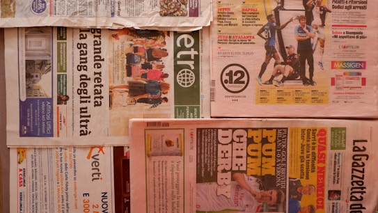 Multiple newspapers are laid out with various headlines and images. The layout includes different sections and pages, with colors ranging from a light tan to vibrant reds and greens. Advertisements and articles are visible with bold headlines and images related to sports and news events. The newspapers are arranged horizontally across a flat surface.