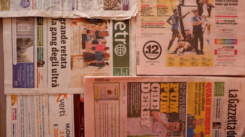 Multiple newspapers are laid out with various headlines and images. The layout includes different sections and pages, with colors ranging from a light tan to vibrant reds and greens. Advertisements and articles are visible with bold headlines and images related to sports and news events. The newspapers are arranged horizontally across a flat surface.
