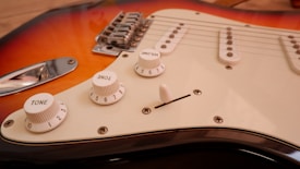 Close-up view of an electric guitar focusing on the control knobs and part of the body. The guitar features a sunburst finish with shades of orange and red, and a white pickguard with three white knobs for tone and volume control. The metal bridge and strings are also visible.