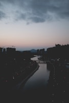 A serene cityscape at dusk with a river flowing through the center. The skyline shows silhouettes of buildings against a soft, pastel-colored sky. Streetlights are seen along the riverbanks, glowing faintly in the fading light.
