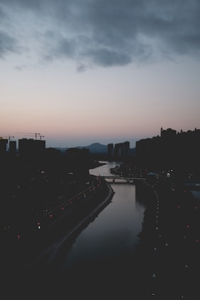 A serene city skyline at dusk with soft, warm lighting highlighting the buildings and a calm river in the foreground.