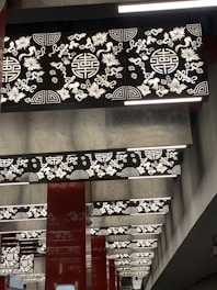 A series of decorative panels on the ceiling feature intricate floral and geometric patterns. The panels are illuminated with light, highlighting the contrast between the ornate designs and the dark background. The columns between panels are painted in a deep red color, contributing to the elaborate and traditional aesthetic.