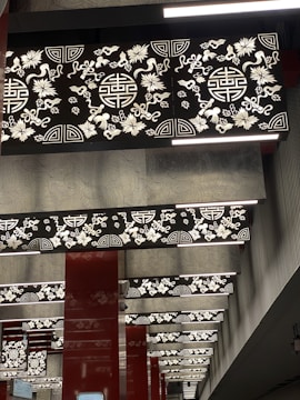A series of decorative panels on the ceiling feature intricate floral and geometric patterns. The panels are illuminated with light, highlighting the contrast between the ornate designs and the dark background. The columns between panels are painted in a deep red color, contributing to the elaborate and traditional aesthetic.