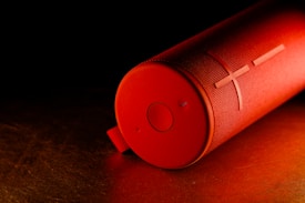 A cylindrical red portable speaker lies on its side against a dark background. The speaker has a textured surface with a prominent plus and circle symbol on one end, along with a fabric loop for carrying.