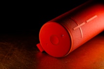 A cylindrical red portable speaker lies on its side against a dark background. The speaker has a textured surface with a prominent plus and circle symbol on one end, along with a fabric loop for carrying.