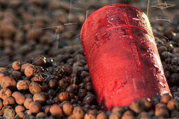 A red portable speaker partially laying on a surface covered with round, brown pebbles and being sprayed with water droplets.