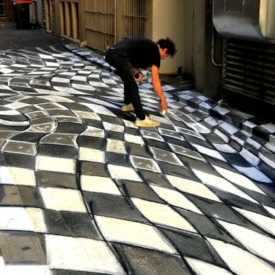 A person is creating a three-dimensional optical illusion on a pavement using paint. The artwork appears to be a black and white checkered pattern that creates a wave-like effect, giving the impression of a distorted or undulating surface.