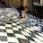 A person is creating a three-dimensional optical illusion on a pavement using paint. The artwork appears to be a black and white checkered pattern that creates a wave-like effect, giving the impression of a distorted or undulating surface.