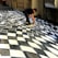 A person is creating a three-dimensional optical illusion on a pavement using paint. The artwork appears to be a black and white checkered pattern that creates a wave-like effect, giving the impression of a distorted or undulating surface.