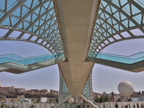 A modern architectural bridge with an arched, lattice-style roof primarily made of glass and metal. The structure spans across from a close viewpoint and leads into the distance. There is an orb-like structure in the background along with a cityscape featuring a few low-rise buildings and trees.