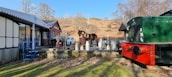 Open-air scene featuring a drinks stand with several milk churns set up on a wooden platform. A metal sculpture of a horse stands as a centerpiece next to a brick building labeled 'Erwood.' A vintage green and red train car is situated on tracks adjacent to the platform. A white car is parked nearby, and there are a few scattered chairs and a table. The surrounding landscape includes bare trees and dry, grassy hills under a clear blue sky.