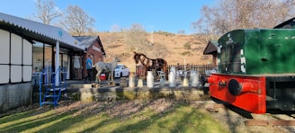 Open-air scene featuring a drinks stand with several milk churns set up on a wooden platform. A metal sculpture of a horse stands as a centerpiece next to a brick building labeled 'Erwood.' A vintage green and red train car is situated on tracks adjacent to the platform. A white car is parked nearby, and there are a few scattered chairs and a table. The surrounding landscape includes bare trees and dry, grassy hills under a clear blue sky.