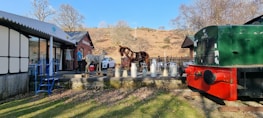 Open-air scene featuring a drinks stand with several milk churns set up on a wooden platform. A metal sculpture of a horse stands as a centerpiece next to a brick building labeled 'Erwood.' A vintage green and red train car is situated on tracks adjacent to the platform. A white car is parked nearby, and there are a few scattered chairs and a table. The surrounding landscape includes bare trees and dry, grassy hills under a clear blue sky.