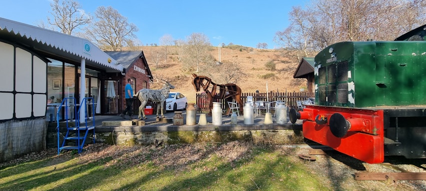 Open-air scene featuring a drinks stand with several milk churns set up on a wooden platform. A metal sculpture of a horse stands as a centerpiece next to a brick building labeled 'Erwood.' A vintage green and red train car is situated on tracks adjacent to the platform. A white car is parked nearby, and there are a few scattered chairs and a table. The surrounding landscape includes bare trees and dry, grassy hills under a clear blue sky.