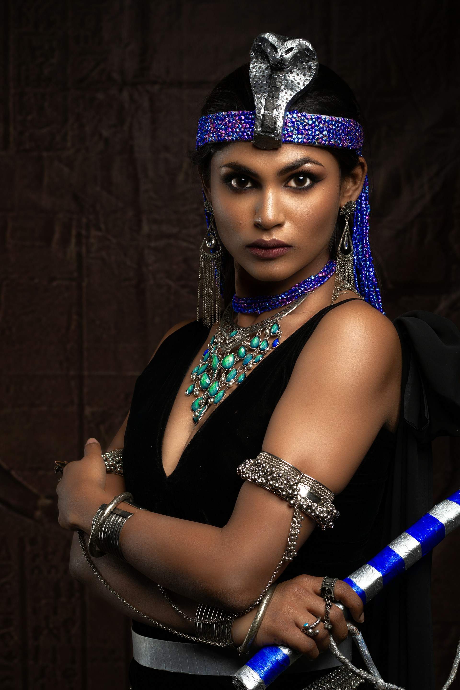 A person standing with a serious expression, adorned in elaborate jewelry and attire resembling ancient Egyptian style. The individual wears a headdress with a snake motif, intricate necklaces, and bold bracelets while holding a staff.