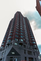 A tall, modern high-rise building with numerous windows and a red-brick facade. The perspective is from the ground looking upwards, showcasing its impressive height against a cloudy sky. The lower portion of the building features a sign reading 'Train More'.