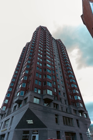 A tall, modern high-rise building with numerous windows and a red-brick facade. The perspective is from the ground looking upwards, showcasing its impressive height against a cloudy sky. The lower portion of the building features a sign reading 'Train More'.