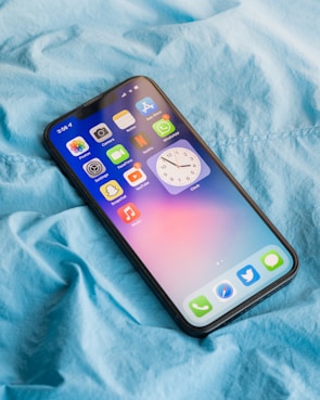 A smartphone displaying several app icons is laying on a wrinkled blue fabric surface. The screen shows apps like Photos, Camera, Notes, and settings, among others. The blue fabric suggests a casual, comfortable setting.
