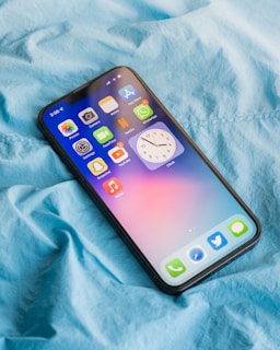 A smartphone displaying several app icons is laying on a wrinkled blue fabric surface. The screen shows apps like Photos, Camera, Notes, and settings, among others. The blue fabric suggests a casual, comfortable setting.