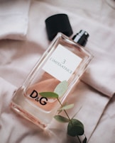 A glass perfume bottle labeled 'L'Imperatrice 3' with a black cap lying next to it, resting on a soft fabric with a small branch of green leaves placed nearby. The branding 'D&G' is prominently displayed on the bottle.