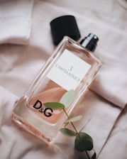 A glass perfume bottle labeled 'L'Imperatrice 3' with a black cap lying next to it, resting on a soft fabric with a small branch of green leaves placed nearby. The branding 'D&G' is prominently displayed on the bottle.