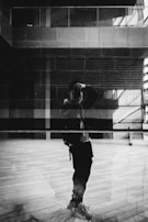 A monochromatic image featuring a reflection of a person taking a photograph in a large glass surface. The background displays an urban setting with architectural elements such as windows and walls, creating a layered effect. The interplay of light and shadows enhances the composition.