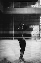 A monochromatic image featuring a reflection of a person taking a photograph in a large glass surface. The background displays an urban setting with architectural elements such as windows and walls, creating a layered effect. The interplay of light and shadows enhances the composition.