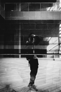 A monochromatic image featuring a reflection of a person taking a photograph in a large glass surface. The background displays an urban setting with architectural elements such as windows and walls, creating a layered effect. The interplay of light and shadows enhances the composition.