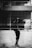 A monochromatic image featuring a reflection of a person taking a photograph in a large glass surface. The background displays an urban setting with architectural elements such as windows and walls, creating a layered effect. The interplay of light and shadows enhances the composition.