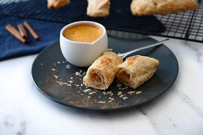 A dark plate features flaky pastry rolls filled with slices of fruit, likely apple. Beside them is a small white cup filled with a creamy, brown drink. Cinnamon sticks rest on a dark blue cloth, and more pastries are on a cooling rack in the background. The marble surface adds a touch of elegance.