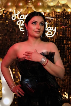 A woman in an elegant black dress stands in front of a decorative background featuring golden balloons and a bright sign with cursive letters. She has a confident expression and wears jewelry including earrings, bracelets, and rings.