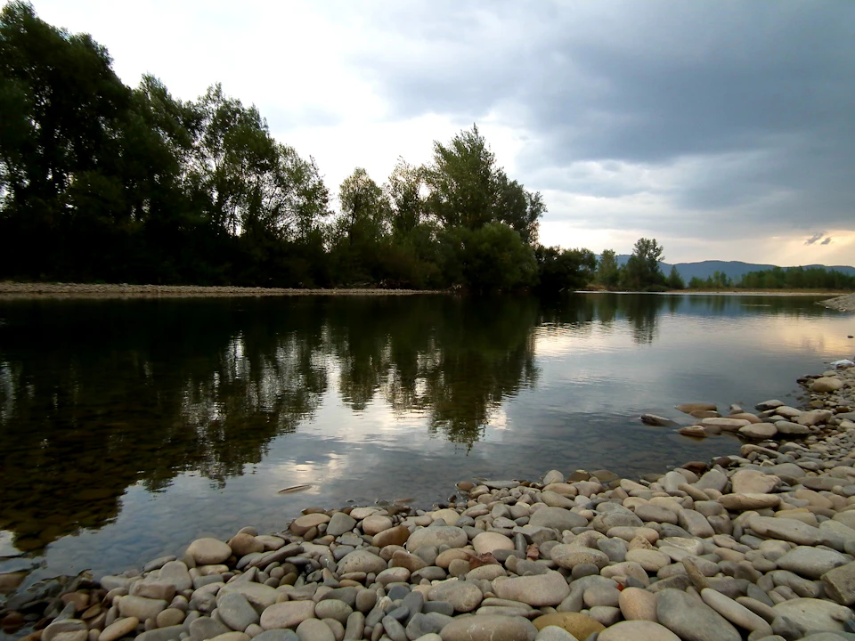 Serene riverbank scene with rocky shore and calm water reflecting overcast sky above landscape