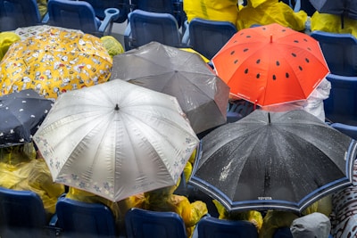 Several colorful umbrellas, including a red one with black spots and a yellow one with cartoon characters, are opened to shield people from rain. Rain droplets are visible on the umbrellas, and the people underneath are wearing yellow raincoats, seated in what appears to be a stadium or outdoor event seating.